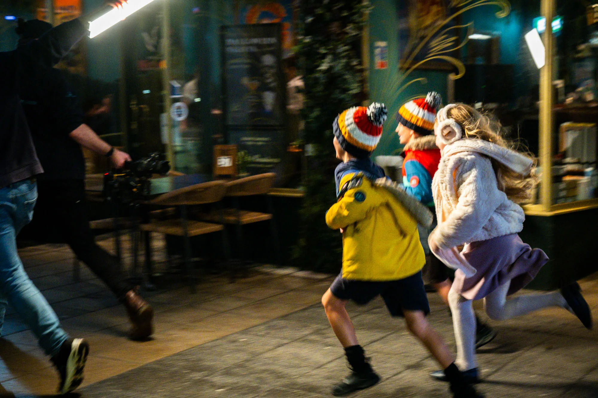 Three children in winter clothing, wearing colourful hats, run energetically on a city street at night. A camera crew follows closely, capturing their joyful motion.